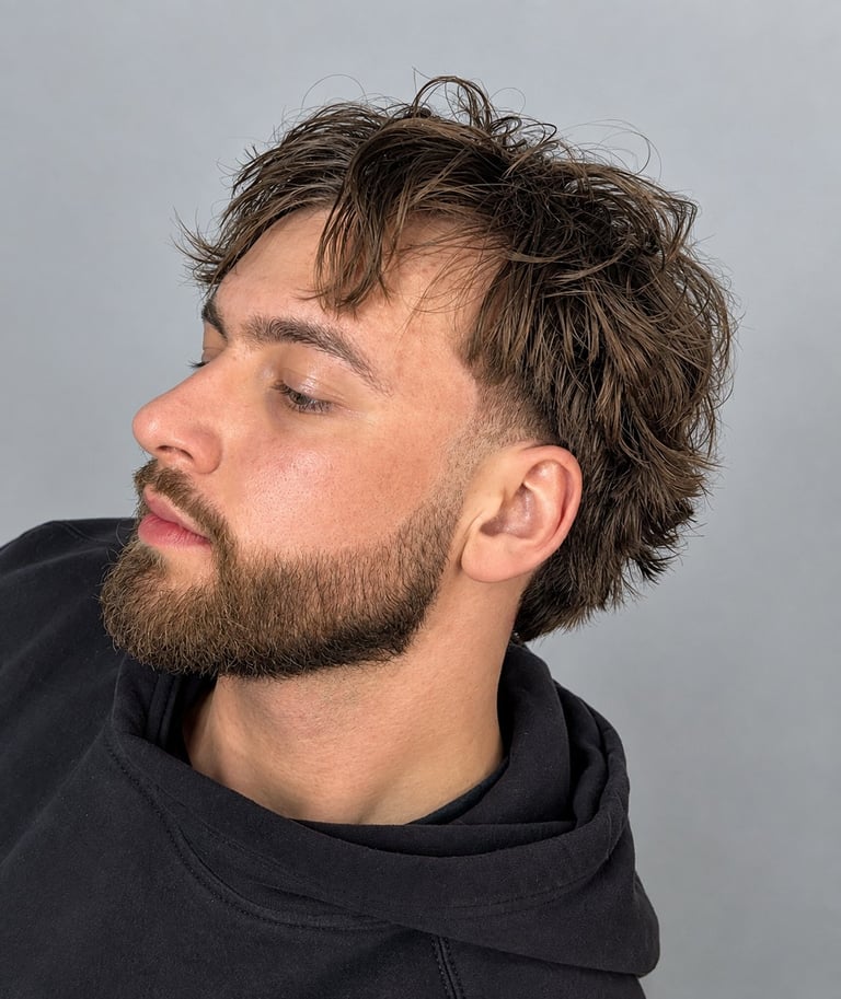 Profile view of a man with dark wavy hair and beard wearing a black hoodie against a gray background