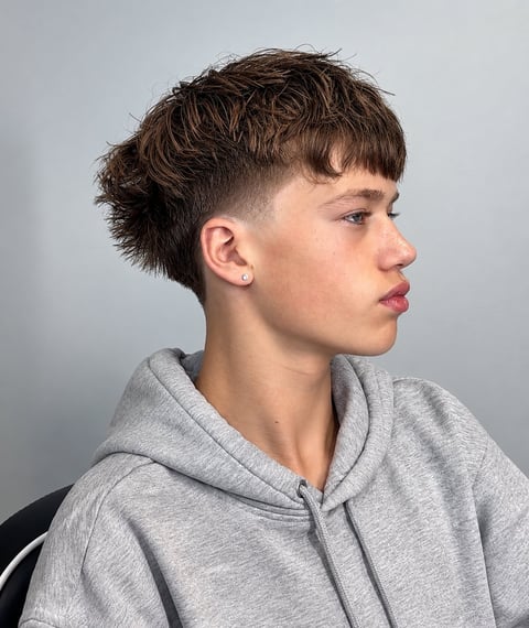 Profile view of a young person with short dark brown textured hair and undercut fade, wearing a light gray hoodie against a neutral gray background