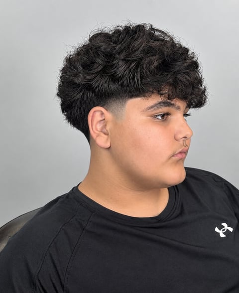 Side profile of a young person with dark curly hair styled with an undercut, wearing a black t-shirt against a gray background