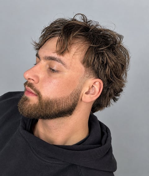 Side profile of a man with dark wavy hair and beard wearing a black hoodie against a gray background