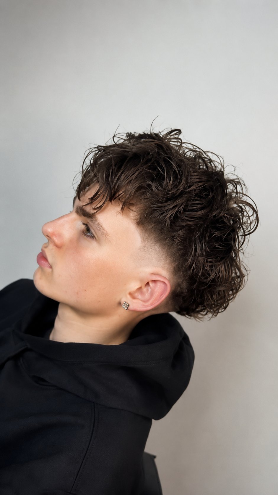 Person in profile wearing black hoodie with curly dark brown hair and silver ear piercing against white background